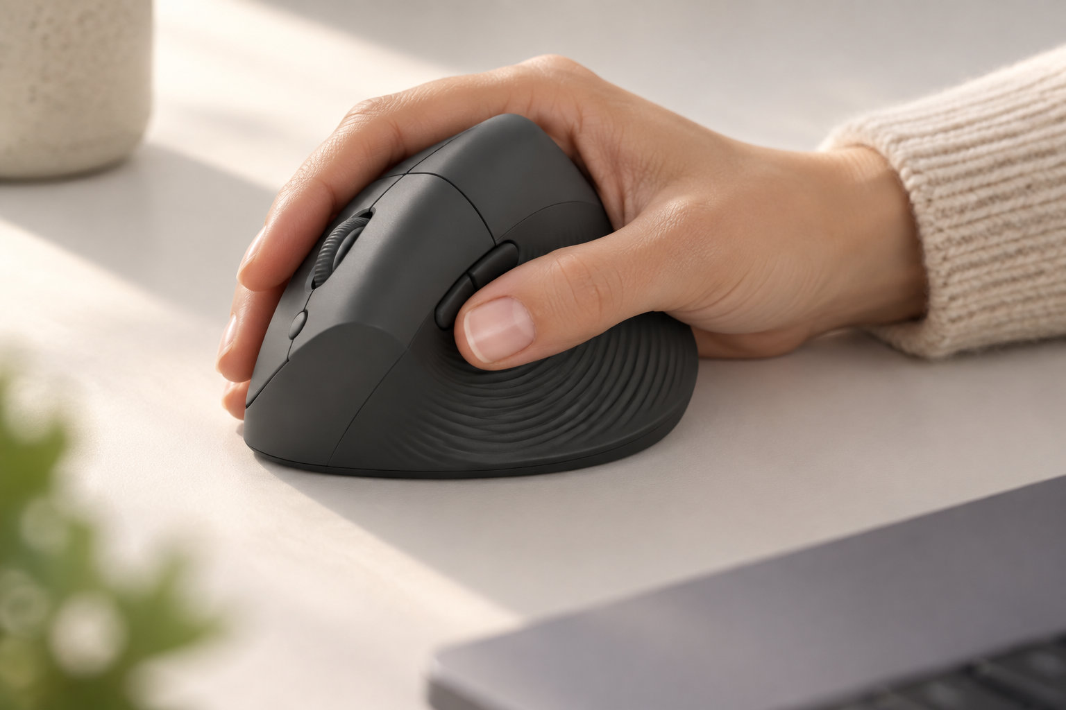 Une main posée sur une souris ergonomique noire sur un bureau clair.