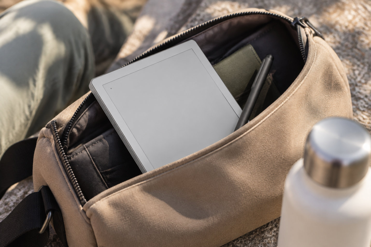 Une sacoche beige ouverte contient une tablette à écran éteint et un stylet, avec une gourde métallique au premier plan.